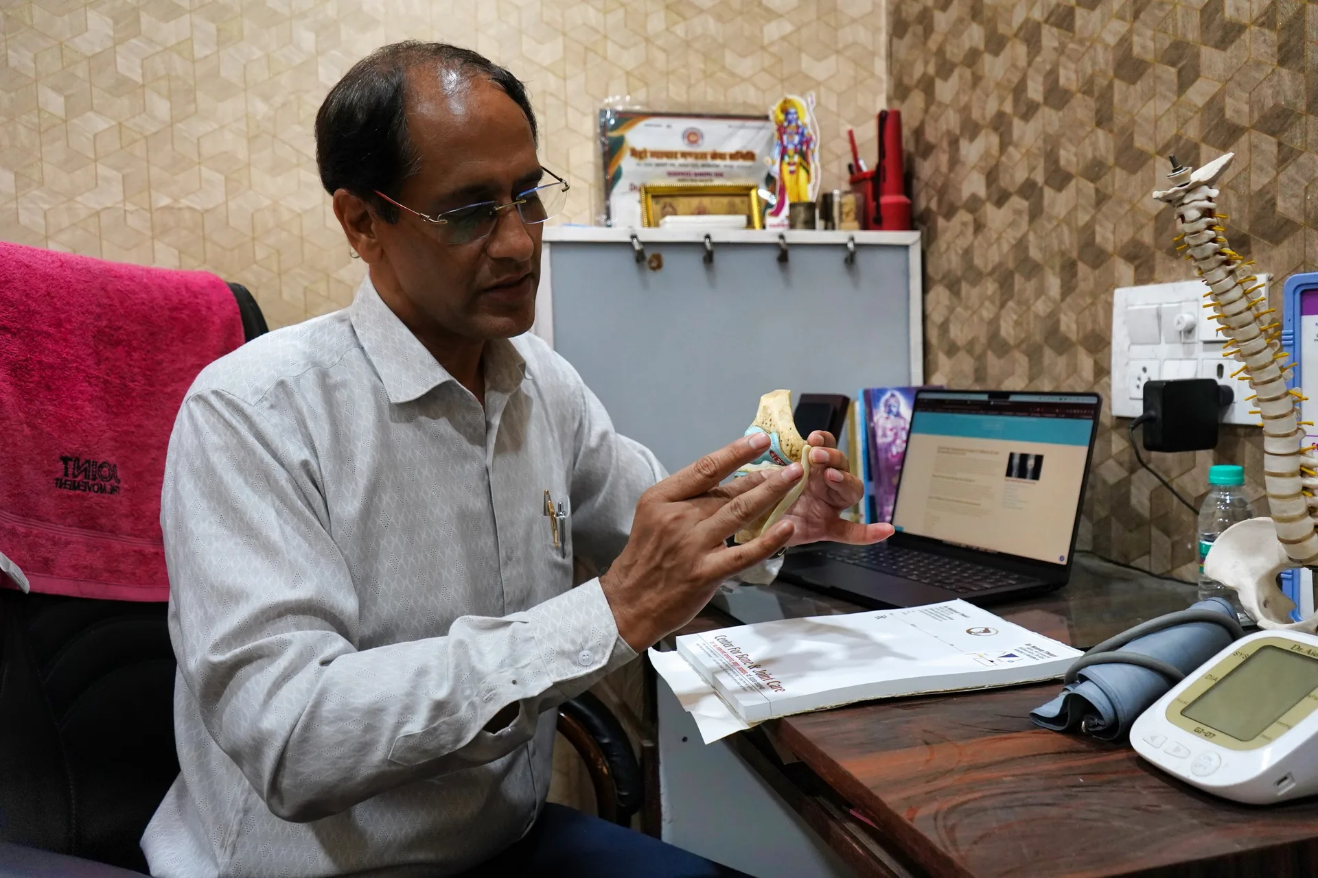 Dr. Mukesh Tiwari explaining with a knee joint model at his clinic desk, seen from the side